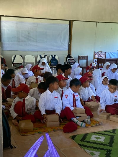 This image depicts a group of Indonesian schoolchildren, likely elementary age, gathered in what appears to be a classroom or community hall in Handil Baru, Indonesia. The children are dressed in white shirts with red ties and red pants or skirts, and some are wearing red caps. They are seated on the floor on mats and some cardboard boxes, with a few chairs and backpacks visible in the background. The walls are light-colored, and there is a large white banner with text on the left side of the image. A sign with the word "SANG" in colorful letters is visible on the right. Many of the children are looking in the same direction, suggesting they are listening to a speaker or watching something. Some children are holding cardboard boxes. The overall atmosphere appears to be one of an educational or community event.