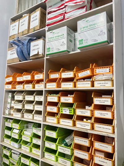 The image shows a storage shelf filled with medical supplies. The shelf is divided into multiple compartments, each holding boxes or containers of various medical items. The items appear to be organized by type and possibly by brand. Many of the containers have labels with text in English, some of which are partially visible. Examples of visible text include "GP Care," "3-Way Stopcock with Extension Tube," "CUTTING," "VIGILENZ," "TRITON," "JARUM," and product codes like "B.SIDE 3-0 5266." There are also labels in Indonesian, such as "URINAL LAKI-LAKI," "URINAL WANITA," "SALTER DEWASA," "SALTER ANAK," and "SALTER BAYI." The setting appears to be a medical storage area or pharmacy, likely in Indonesia given the Indonesian text. The lighting suggests an indoor environment. There are no people visible in the image, and the focus is solely on the organized medical supplies.