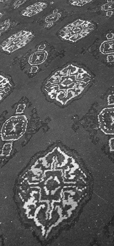 The image is a close-up, angled shot of a carpet with a repeating geometric pattern. The carpet is dark with lighter colored intricate designs that resemble paisley or floral motifs. The lighting is even, and the image is in black and white, suggesting a focus on texture and pattern rather than color. There are no people or specific objects identifiable within the frame. The environment appears to be an indoor space, as indicated by the presence of the carpet. The location is identified as Yazy, Turkmenistan.