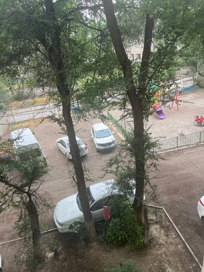In an outdoor courtyard in the city of Shu, Kazakhstan, several white vehicles are parked on a dirt and asphalt surface. A white cargo van is positioned to the left, angled towards the viewer. To its right, a white sedan is parked. Further to the right, another white sedan is visible. In the foreground and scattered throughout the scene are several trees with dense green foliage. Beyond the vehicles, a playground area is present, featuring a purple slide, a red merry-go-round with horse-shaped seats, and other playground equipment. A metal fence encloses part of the playground. Buildings can be seen in the background, with some evidence of construction or repair. The ground is a mix of dirt, gravel, and paved areas, with some concrete curbs visible.
