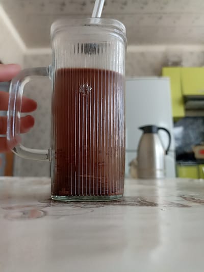 A close-up, low-angle shot captures a glass mug filled with a dark brown liquid, likely chocolate milk or coffee, resting on a patterned surface that appears to be a tablecloth or countertop. A hand is visible on the left, holding the mug's handle. The mug itself is made of clear, fluted glass and contains a white straw. In the background, a silver thermos with a black lid stands on a white surface, possibly a refrigerator. The setting appears to be a kitchen, with hints of lime green cabinetry and a white appliance visible behind the thermos. The image is focused on the mug and the immediate foreground, with the background out of focus. The overall scene suggests a casual, domestic environment, possibly in a home in Кош-Дөбө, Kyrgyzstan, given the prompt's contextual information.