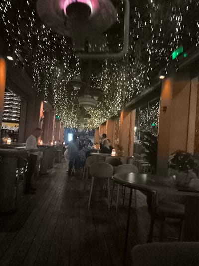 This low-light photograph captures the interior of a modern restaurant or bar during the evening. The most striking feature is the ceiling, densely covered with numerous small, bright string lights that create a sparkling, "starry night" ambiance. Several larger, dark, bowl-shaped pendant lights also hang from above. In the foreground, a prominent dark fixture, possibly an industrial-style heater or lamp, emits a distinct purple-pink glow from its core.

The setting includes a dark wooden floor and various seating arrangements. On the left, a bar area is visible with shelves displaying bottles, and a man in a light-colored shirt stands near the counter. Numerous patrons are scattered throughout the mid-ground, seated or standing around round tables with upholstered chairs and stools, engaged in socializing. Small, warm-toned lamps or candles provide accent lighting on the tables.

The right side of the space features large glass panels or windows, through which faint outdoor lights and some plants are discernible, suggesting a view to an exterior area or a covered patio. Warm, orange-toned architectural pillars or wall sections line this side. Small green rectangular lights are mounted on the upper wall or ceiling, possibly serving as indicators or exit signs. The overall atmosphere is relaxed and social, characteristic of an evening gathering in a contemporary establishment. No discernible text is visible in the image.