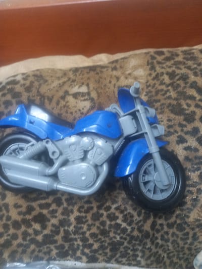 A toy motorcycle is displayed on a leopard-print surface, positioned on the right side of the frame. The motorcycle is primarily blue and gray, featuring a detailed engine and exhaust system. A wooden surface is visible at the top of the image. The environment does not provide any information about the city of Maloye Lugovoye, Russia.
