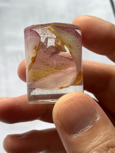 The image presents a close-up view of a person's hand holding a small, clear, rectangular object. The hand, with visible skin texture on the thumb and parts of other fingers, holds the object horizontally in the center of the frame. The fingernails are clean and show some natural ridges.

The object is transparent, likely made of resin or glass, and contains various dried flower petals. These petals are predominantly light purple and pink, with some sections displaying yellowish-brown tones, indicating the natural variations or dried state of the plant material. Numerous tiny air bubbles are trapped within the clear material, creating a speckled or effervescent effect throughout the object. The object's edges appear to be slightly rounded.

The background is out of focus and appears to be a plain, light-colored surface, possibly suggesting an indoor setting. The lighting is bright and even. The activity captured is simply the act of holding and presenting this decorative item. No specific location cues, time of day, weather conditions, or emotions are discernible. There is no visible text in the image.
