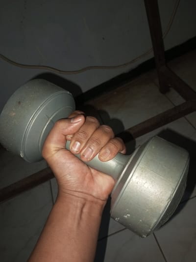 A hand grips a grey, dumbbell. The skin tone of the hand is dark. The nails are light in color. The dumbbell has a cylindrical shape with larger, rounded ends. A power cord stretches along the wall behind the dumbbell. A portion of a metal chair can also be seen behind the dumbbell. The flooring is tiled. The lighting is dim.