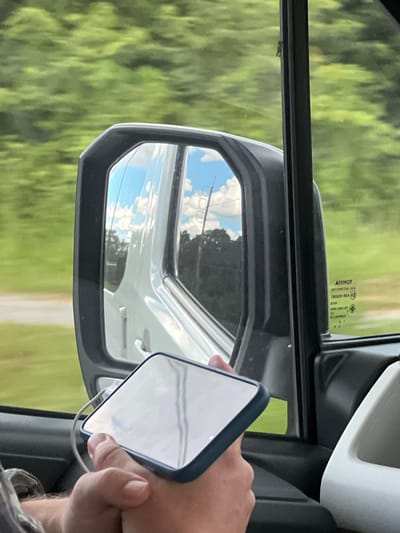 The image shows a person's hand holding a smartphone, reflecting the sky and clouds in its screen. The hand is positioned near a car's side mirror, which also reflects the sky and clouds, as well as a blurry green landscape outside the vehicle. The scene appears to be from inside a car, possibly in motion, given the reflection and the context of the side mirror. The weather seems to be clear with scattered clouds. The greenery suggests a rural or natural setting. There is some visible text on the car window, though it is mostly illegible in the provided image. The overall impression is a casual, everyday moment captured while traveling. The visible text on the car window, near the mirror, includes "ATOYOT," "E4," and a series of numbers and letters that are difficult to decipher clearly. The location is given as Ban Ko Pru, Thailand.