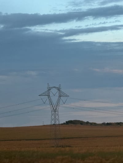 The image shows a tall, metallic electricity pylon standing in a field of dry, golden grass under a cloudy blue sky. The pylon is the central focus, its lattice structure clearly visible. Several power lines extend horizontally from the pylon across the frame. In the background, a gentle slope covered with sparse green foliage forms the horizon line, with a few scattered trees on top. The sky is filled with horizontal bands of light and dark gray clouds, suggesting an overcast or twilight condition. The image appears to have been taken from a low angle, emphasizing the height of the pylon and the vastness of the sky. The scene is devoid of any human presence or activity. The location is identified as Krustina, Bulgaria.