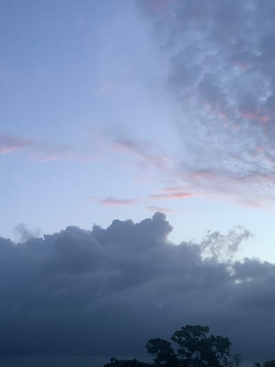 The image depicts a sky with clouds and a hint of tree foliage at the bottom. The sky transitions from a pale blue at the top to a darker, more ominous gray towards the bottom. Several cloud formations are visible, ranging from wispy, pink-tinged streaks in the upper portion to dense, dark gray masses lower down. The pink hues suggest the presence of twilight. A small portion of dark green tree leaves is visible at the very bottom edge of the frame, indicating an outdoor, potentially natural setting. The scene is static and devoid of human presence or activity. The location is identified as Karandugoda, Sri Lanka, implying this is a view of the sky in that region.