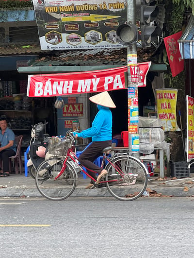 A woman wearing a conical hat and a blue long-sleeved shirt is riding a red bicycle with a basket on the front. The bicycle is on a paved street in front of a shop. The shop has a red banner with Vietnamese writing that says "BÁNH MỲ PA". There are also several posters and signs on the wall of the shop. A man is sitting on a chair to the left of the shop. A scooter is parked next to the bicycle. The environment appears to be a street in a city in Vietnam, likely Tân Quang Commune, based on the signage. The overall scene is a snapshot of daily life on a street in Vietnam.