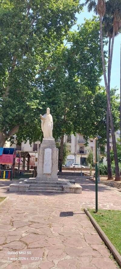 The image depicts a statue in Canals, Spain. The statue is light-colored and stands atop a stone base with steps leading up to it. A plaque is visible on the statue's base. Lush green trees surround the statue, providing a backdrop of foliage. Palm trees are also visible, adding to the outdoor setting. A walkway paved with irregularly shaped stones leads away from the statue. In the background, buildings and a glimpse of a playground can be seen.