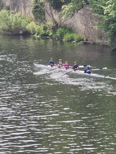 A horizontally oriented photo shows a rowing team of four people in a white boat on a river, likely the River Wear in Durham, United Kingdom, based on the stone embankment on the left side of the image. The river appears to be green with numerous reflections of light across its surface. The team members are positioned one behind the other in the boat and are using oars to propel themselves through the water. The embankment features green vegetation and stone wall. The sky is not visible but it is likely that this photo was taken during the day because of the brightness of the photo.