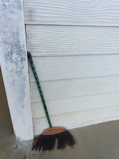 A dark green broom with a light green leaf pattern on its handle is depicted leaning against a building structure in Man Gyat, Myanmar. The broom's black synthetic bristles and orange-brown plastic head rest on a grey concrete surface, with minor debris visible near the bristles. The broom is positioned at the junction of two vertical surfaces: a distressed, light grey pillar or trim on the left, exhibiting peeling paint and a rough texture, and a white horizontal siding on the right, featuring a wood grain pattern and secured with visible screws. A black loop is present at the top of the broom's handle. No individuals are present in the image.