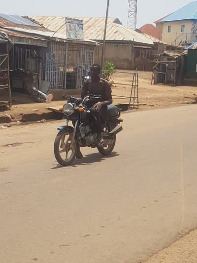 In Tukur Tukur, Nigeria, a Black man is riding a blue and silver motorcycle on a paved road. He is wearing a dark-colored shirt and dark pants. The motorcycle has a license plate that is partially visible. In the background, there are several buildings with corrugated metal roofs, some of which appear to be shops or small businesses. One building has solar panels on its roof. A tall telecommunications tower is visible in the distance. The environment appears to be an urban or semi-urban area with a dry, dusty atmosphere.