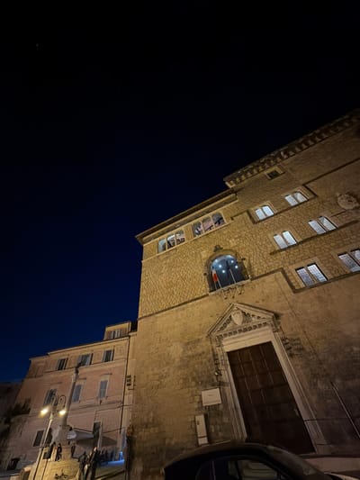 The photograph depicts a night scene in Tarquinia, Italy. A tall, multi-story stone building with arched windows and a large wooden door dominates the right side of the frame. A flag is visible through one of the arched windows. To the right of the building, the rear of a vehicle is partially visible, suggesting a street or road. Below the tall building is a smaller, lighter-colored building with multiple windows. Streetlights are visible, illuminating the facades of the buildings. The left side of the frame is dominated by a dark blue night sky.