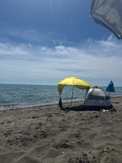 An outdoor, eye-level shot captures a beach scene on a sunny day in Aheloy, Bulgaria. In the foreground, dark sand is disturbed by ripples and tracks. Beyond the sand, the blue-green sea meets the shore. A few umbrellas and a tent are set up on the beach. A bright yellow umbrella is positioned over a grey and black tent. To the right of the tent, a blue umbrella stands upright. In the background, the horizon separates the sea from a hazy sky with scattered white clouds and contrails from an aircraft. The scene suggests a day at the beach with some beach gear.
