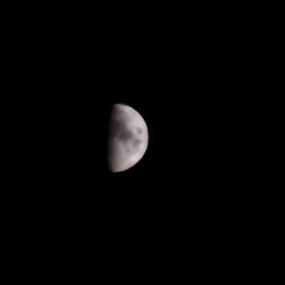 The image shows a partial view of the moon against a black sky. The moon is illuminated on the right side, appearing as a waxing gibbous or waning gibbous phase. Darker patches, representing maria or craters, are visible on the illuminated surface. The image is out of focus, giving the moon a soft, blurred appearance. The background is uniformly black, indicating it is a night sky with no other celestial bodies or light pollution visible. There are no elements in the image that suggest a specific location in El Perelló, Spain, or any urban environment.
