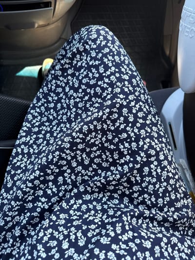 The image shows a close-up view of a person's legs wearing a dark blue dress with a white floral pattern. The person is sitting in a vehicle, as indicated by the car interior visible in the background, including a gray dashboard and black floor mats. Sunlight is entering the car from the left, casting a shadow on the floor. To the right, a portion of a white object, possibly a car seat or a container, is visible with text that appears to say "SIPS PROTEC." The image does not provide any specific information to locate it in Volpertshausen, Germany.
