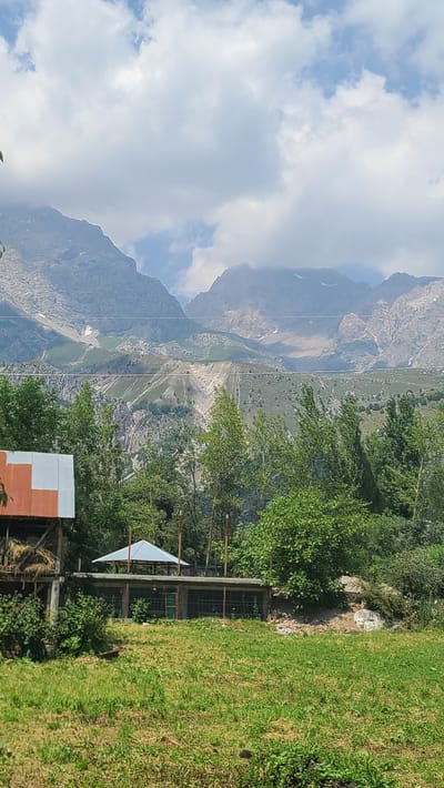 The image depicts a mountainous landscape in Arstanbap, Kyrgyzstan. In the foreground, a grassy field leads to a series of structures. To the left, a part of a building with a red and white corrugated metal roof is visible, with hay bales stacked beneath it. Adjacent to this, a long, low structure with an open roof section covered by a white canopy extends horizontally. This structure appears to be an enclosure, possibly for animals, with wire mesh sides and wooden posts.
Behind these structures, a dense line of green trees forms a natural barrier. Beyond the trees, the terrain rises dramatically, revealing steep, rocky mountainsides with patches of green vegetation and snow-capped peaks under a cloudy sky. Power lines are visible crisscrossing the middle ground, stretching across the mountains. The overall scene suggests a rural setting nestled within a grand natural environment. No people are visible in the image.