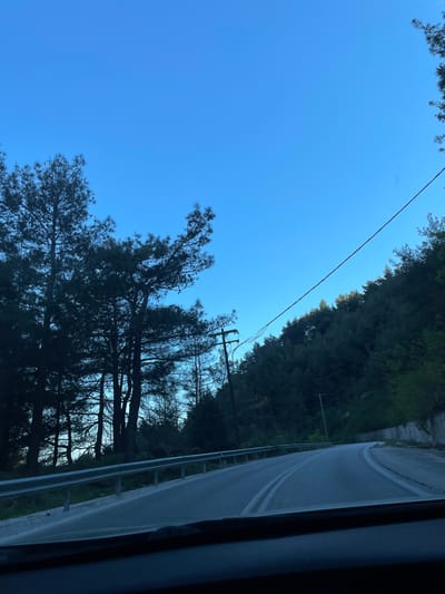The image, rotated counter-clockwise, shows a view from a vehicle traveling along a road in the Stavroupoli Municipal Unit, Greece. The sky is clear and blue. A road with double yellow lines runs along the right side of the frame, bordered by a metal guardrail. Trees are visible on both sides of the road, denser on the right. A utility pole with wires is also visible.