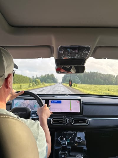 The image shows the interior of a car being driven by a person wearing a cap. The car is on a paved road, which appears to be a highway, in a rural area with green fields and trees on either side. The sky is overcast. The car's dashboard displays navigation information, showing a route to Яган-Докья, Russia, with 4.8 km remaining. The car appears to be moving, as suggested by the motion blur in the background and the driver's hands on the steering wheel. There are no other people visible in the car.