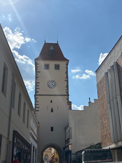 The image shows the historic Mělník Castle Tower in Mělník, Czechia, under a bright blue sky with scattered white clouds and strong sunlight. The tower, a prominent landmark, features a red tiled roof with a small green turret and two spires. Its cream-colored facade is adorned with a large clock face with Roman numerals and a pointed archway at its base, through which a street scene is visible. The tower is flanked by buildings on either side: a lighter colored building with windows on the left and a building with a reddish-brown tiled facade and modern vertical elements on the right.
In the foreground on the left, a man in a red shirt stands outside a shop with the signage "STUDIO" and "ARTE PRO". The street appears to be paved. Further into the arched entrance of the tower, people can be seen walking, suggesting the passage is open to the public. A truck is parked in the lower right portion of the frame, partially obscuring the view of the building to the right. The overall scene captures a sunny day in a historic European city.