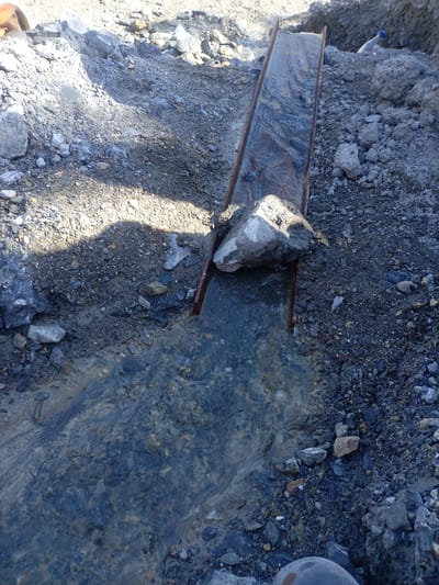 This is a close-up, high-angle shot of a makeshift sluice box, likely used for gold panning or mineral extraction, set up in a rough, rocky terrain. The sluice is made of a long, narrow channel with reddish-brown wooden or metal sides, through which water flows. The water is clear in places, showing pebbles and sand beneath the surface, and murky in others where it carries sediment. Large grey rocks and smaller gravelly debris surround the sluice, indicating a natural or disturbed landscape. A large grey rock is perched on the edge of the sluice at the top of the frame. The lighting suggests daylight, with shadows cast on the ground. The scene appears to be located outdoors, possibly in a remote area. The context of Zeravshan-2, Tajikistan, suggests this could be related to mining or prospecting activities in that region. There are no people visible in this image.