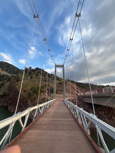 This image depicts a suspension bridge in Ait Mazigh, Morocco. The bridge has a wooden plank walkway with white metal railings and support cables. The view is from the bridge itself, looking towards one of the anchor points. The landscape features rocky hills with sparse vegetation on both sides of the bridge. The sky is partially cloudy, with a mix of blue and gray clouds. Another, larger bridge structure is visible at the far end of the visible suspension bridge. A body of water is partially visible below the bridge. A portion of a person's finger is visible in the lower right corner of the image.