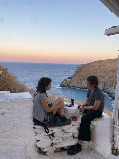 The image displays two individuals seated on a whitewashed stone structure overlooking the Aegean Sea and a rugged, arid coastline, consistent with a location such as Απολλωνία, Greece. The scene is illuminated by the soft light of sunset or dusk, evidenced by the sky's gradient from light blue overhead to orange and pink at the horizon.
On the left, a female with dark, wavy hair wears a grey hooded t-shirt, dark shorts, and dark robust shoes. She has hoop earrings and visible tattoos on her left lower leg. Her body is angled towards the male, suggesting engagement in conversation. On the right, a male with curly brown hair wears a grey t-shirt, dark trousers, and dark shoes. He is also oriented towards the female.
Between the two individuals, a small, whitewashed stone pedestal serves as a surface for various objects: a clear plastic cup containing a light-colored drink with a lime slice, a purple energy drink can, and a small, dark object. Near the male, a second clear plastic cup holds what appears to be a green plant or herbs. A dark bag hangs from the stone structure near the female's feet. The immediate foreground features the rough, whitewashed stone construction. In the background, the deep blue sea meets the base of steep, barren cliffs and headlands. A portion of a weathered, whitewashed wooden support beam is visible on the far right.