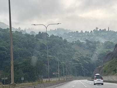 The image depicts a highway scene in Tsakva, Georgia, under an overcast sky. A light-colored car with a roof box is driving on the right lane of the multi-lane road. The highway is bordered by a concrete barrier on the right and a steep, tree-covered mountainside on the left, with a prominent rock face. Numerous lampposts line the left side of the road. In the distance, rolling hills covered in dense green foliage are visible, with scattered houses and trees dotting the landscape. The atmosphere appears hazy or foggy, obscuring the finer details of the distant scenery. A single lamppost stands in the foreground on the left side of the image, appearing to be a prominent feature of the roadside infrastructure.
