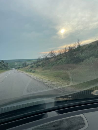 The image depicts a scenic view from a vehicle, likely a car, as evidenced by the dashboard and speaker visible on the right side of the frame. The perspective is from inside, looking out through the windshield.

The primary subjects are a landscape and the sky. A winding asphalt road is seen in the foreground, curving to the right. To the left of the road, a steep embankment covered with sparse vegetation and bare trees rises. Beyond the embankment, a vast, flat landscape stretches to the horizon, dotted with agricultural fields in various shades of green.

The sky is overcast with diffused sunlight, suggesting it is either early morning or late afternoon. The sun is a bright, hazy orb visible in the upper left quadrant of the image. The weather appears to be calm, with no strong winds or precipitation indicated.

Notable details include the reflections on the windshield, which obscure parts of the landscape and create a layered effect. The texture of the road surface and the details of the vegetation on the embankment are discernible. The presence of a small white car is visible in the distance on the road.

There is no visible text in the image. The overall mood conveyed is one of travel and contemplation of a natural, somewhat muted landscape. The location is identified as Tartaul de Salcie, Moldova.