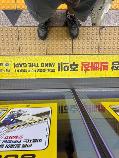 An overhead photograph captures the lower legs and feet of an individual positioned on a subway platform in Jongno-gu, Seoul, South Korea. The person wears light-colored shorts, black and white striped socks, and dark grey athletic shoes. A white bag with printed text is visible near their right foot. The platform features light grey speckled surfacing and bright yellow tactile paving with raised circular dots. A prominent yellow warning line spans horizontally across the platform, displaying inverted black Korean characters, including "조심" (caution), and "MIND THE GAP!" in English, along with small Seoul Subway and IBK logos. Below this warning line, a narrow dark grey strip delineates the platform edge, followed by a small gap. Beyond this gap, a reflective surface, consistent with the exterior of a subway train or platform screen doors, displays reflections of yellow advertising panels with cartoon graphics and Korean text, along with a distinct vertical dark line.
