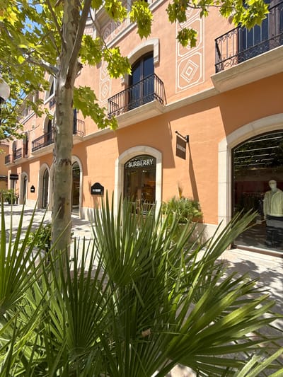 This media file captures an exterior view of a shopping area, likely a designer outlet mall, in la Roca del Vallès, Spain. The scene is dominated by the architecture of the buildings, which have a warm, peachy-orange stucco facade. One building features decorative geometric patterns in white against the stucco. Several windows and balconies with ornate black iron railings are visible.

In the foreground, a large tree trunk and lush green foliage from palm trees partially obscure the view of the buildings. The sunlight suggests it is daytime, and the bright shadows indicate clear weather.

Text visible on the buildings includes "BURBERRY" on a dark sign hanging from the wall of one establishment, and "BURBERRY" above the arched entrance of another shop. A mannequin is visible in the window of one of the stores, dressed in a light-colored garment, suggesting retail activity. The overall impression is one of a pleasant, sunny day in a high-end outdoor shopping environment.