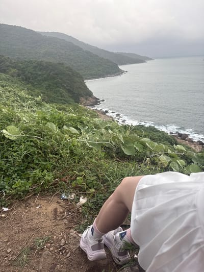 The image displays a coastal viewpoint in Sơn Trà Ward, Vietnam, under an overcast sky. In the foreground, the lower legs of an individual are visible, clad in a white pleated skirt or dress, light pink crew socks, and white and light purple athletic sneakers. The individual is positioned on a dirt-covered incline interspersed with dense green vegetation, with some scattered litter present on the ground. Beyond this, a series of lush, tree-covered hills slope steeply down to a rocky coastline. The sea, appearing grey and slightly choppy, extends into the distance, following the curve of the coastline. No other people or explicit actions/interactions are observed.