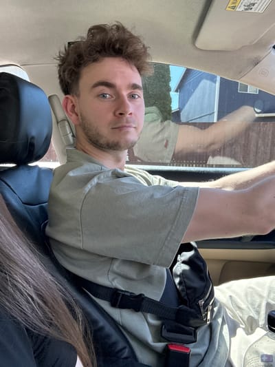 A young Caucasian man with curly brown hair and blue eyes is driving a car in South Hill, United States of America. He is wearing a light green t-shirt and a black fanny pack around his waist. A black leather seat and a tan dashboard are visible. Through the window, a house and trees can be seen.