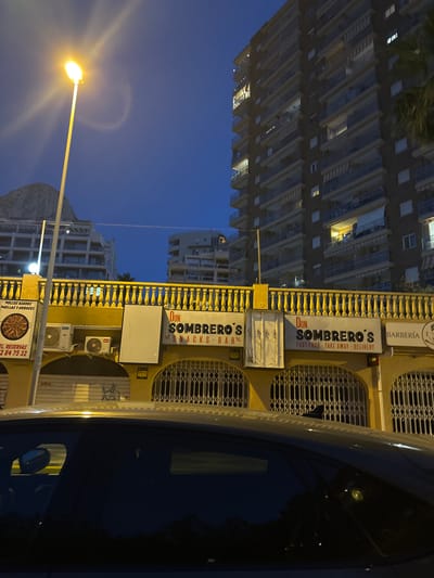 This is a vertically oriented, nighttime photograph taken in Calp, Spain. The scene features the exterior of a building with several signs, a street lamp, and a car. The building has a yellow facade with decorative railings and is situated next to a tall apartment building. One prominent sign reads "DON SOMBRERO'S SNACKS-BAR" with smaller text indicating "FAST FOOD - TAKE AWAY - DELIVERY". Another sign above it says "BARBERÍA" and a sign lower down says "POLLOS ASADOS PAELLAS Y ARROCES" along with a phone number. The street lamp in the foreground emits a bright, warm light. The sky is a deep blue, suggesting it is twilight or early night. A portion of the car's dark blue surface is visible on the right side of the frame, reflecting light from the surroundings. The overall impression is of a commercial street in a coastal town, likely a tourist area, given the presence of a snack bar and barber shop.