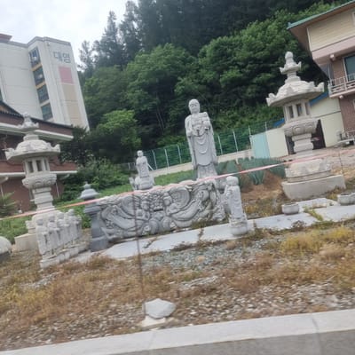 The image captures an outdoor scene featuring several stone statues and a building. In the foreground, a large carved stone relief depicts figures and mythical creatures. To the left of the relief, there are several smaller stone statues of children or monks. In the middle ground, a tall stone statue of a seated Buddha holding a child stands prominently. To the right of the Buddha statue, there is an ornate, multi-tiered stone lantern. Further to the right, part of a brick building with balconies is visible. Behind the statues, a dense forest of trees covers a hillside. In the background, a tall, light-colored building with balconies and Korean text that reads "대영" is visible. The ground is covered with gravel and dry vegetation. The sky is overcast. This scene is likely located in or near Jinbu-myeon, South Korea, given the Korean text on the building and the architectural style.