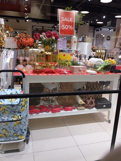 This is a photograph of a store window display in Wołomin, Poland. The display features a white table laden with various decorative items. On the table, there are several red lobster-shaped decorations, a pink jewelry box, white ceramic birds, a white seashell-shaped object, a glass vase with yellow tulips, and a black tissue box with a "Meow" label. A red and white patterned runner with "%" symbols covers the front of the table. Behind the table, there are more decorative items, including gold vases and pink and red flowers. A red "SALE -50%" sign is visible in the upper right portion of the display. To the left of the table, a rolling shopping cart with a blue and yellow lemon-patterned bag is positioned. The floor is tiled in a light beige color. Reflections of lights and interior store elements are visible in the glass.