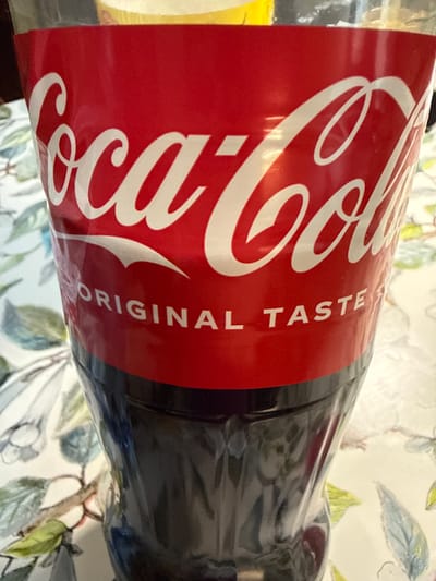A close-up, eye-level shot shows the iconic red label of a Coca-Cola bottle, featuring the white script logo and the words "ORIGINAL TASTE" printed below it. The bottle is positioned on a patterned tablecloth with a floral design in muted greens, blues, and pinks. The upper portion of the bottle, containing a yellow liquid and possibly ice, is slightly out of focus, while the lower, dark liquid-filled section is also somewhat blurred. The scene does not depict any specific location in Borgosatollo, Italy; it is a generic product shot.