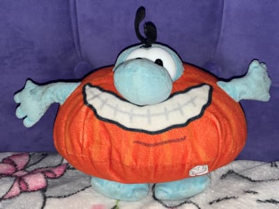 This is a close-up, eye-level shot of a plush toy, likely a character from a children's show. The toy is predominantly orange and light blue, with a large, round orange body that resembles a pumpkin. It has large, white, cartoonish eyes with black pupils, and a prominent light blue nose. A wide, black-lined smile with white teeth is stitched onto the orange body. The toy has light blue arms and hands that are spread wide, and light blue feet. A single black, antenna-like tuft sprouts from the top of its head.
The plush toy is placed on a textured, light-colored surface, possibly a blanket or rug, which has a pattern of pink and black floral designs. In the background, a textured, deep purple surface is visible, likely upholstery from a couch or chair.
The lighting is somewhat dim, creating soft shadows and highlighting the plush texture of the toy and the background. No specific time of day or weather conditions can be inferred. The overall mood is playful and whimsical, typical of children's toys. There is a small white sticker on the orange body of the toy, featuring a cartoonish rabbit face, but no other discernible text is visible in the image. The location context provided, Гаранскі сельскі Савет, Belarus, suggests an indoor setting, possibly a home.