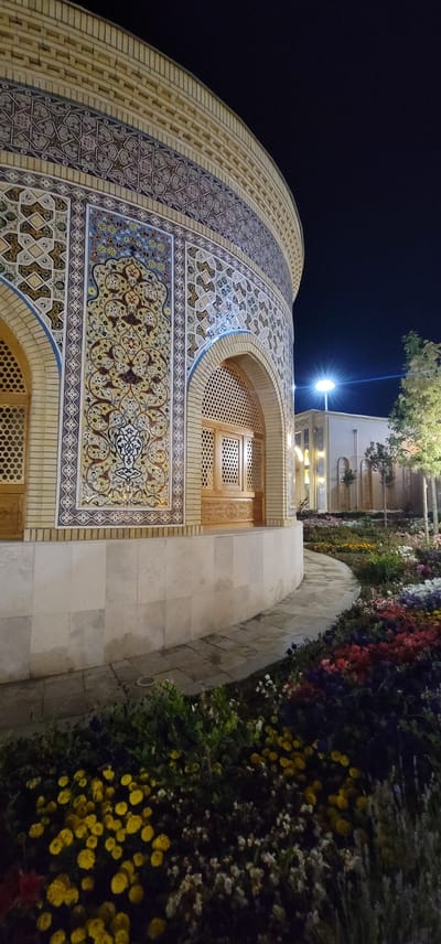 The image shows a night view of a building with intricate tilework, located in the city of Satkak, Uzbekistan. The building features a curved facade adorned with colorful geometric and floral patterns. There are arched openings with latticed wooden windows. In front of the building, a curved pathway winds through a garden filled with a variety of blooming flowers in shades of yellow, red, purple, and white. The background shows another building illuminated by artificial lights, and a tall pole with a bright light fixture. A tree with green foliage stands to the right of the building. The sky is dark, indicating nighttime. There are no people visible in the image.