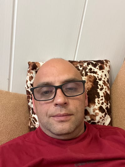A middle-aged bald man wearing glasses and a red shirt is shown in a selfie. He is sitting on a sofa with a cow-print pillow behind his head. The background is a white wall with vertical paneling. The image appears to have been taken indoors.