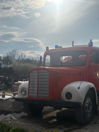 The image shows the front of a vintage, red fire truck with white fenders parked in an outdoor location. The truck features a prominent grill, blue emergency lights on the roof, and a pair of side view mirrors. In the background, trees, headstones, and a building are visible, indicating that the truck is parked near a cemetery in Hrvatska Dubica, Croatia. The sky is partly cloudy with the sun shining brightly. Some building materials and plastic sheets are scattered in the foreground near the truck.