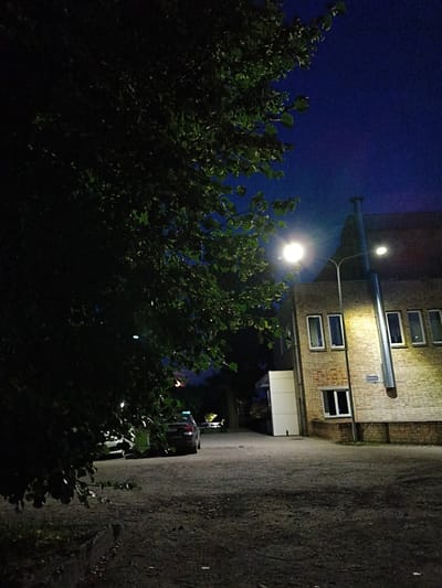 The image shows a nighttime scene in Tukums, Latvia. A large tree with dark green leaves dominates the left side of the frame. In the background, a brick building with multiple windows is illuminated by streetlights. A large, silver vent pipe runs vertically up the side of the building. A streetlight with a bright light is visible on the left, and another dimmer streetlight is on the right. There are two cars parked in the gravel area in front of the building. The sky is a deep blue, indicating dusk or night.