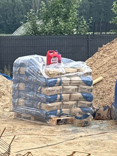 In Opatów, Poland, a construction site is depicted. A large stack of cement bags, wrapped in clear plastic, rests on a wooden pallet. A red jerrycan and a blue bottle are placed on top of the stacked bags. To the right of the cement bags, a pile of sand is visible, with a wooden plank partially embedded in it. In the foreground, rebar from a foundation is scattered on the ground, suggesting ongoing construction work. A dark fence forms the backdrop of the scene, with lush green trees and foliage behind it. The ground appears to be dirt and sand. No people are present in the image.