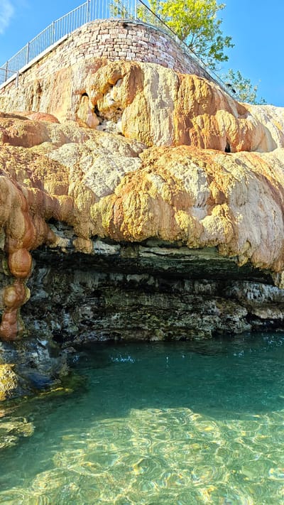 This image captures a scenic view of a mineral formation with cascading water at Loutra Aidipsos, Greece. The formation is composed of layered, textured rock in shades of orange, yellow, and white, suggesting mineral deposits from thermal springs. The rock faces are irregular and appear to have been shaped by water flow over time, with some areas showing stalactite-like formations. Below the mineral deposit, a dark, layered rock face is visible, creating a distinct geological strata.
The lower part of the image features a body of clear, turquoise water. The water's surface is dynamic, with ripples and reflections indicating movement, and small droplets of water are seen falling from the rock face above into the pool. The sunlight illuminates the water, revealing the textured bottom and the clarity of the water.
Above the mineral formation, a low stone wall with a metal railing is visible, suggesting a maintained pathway or viewing area for visitors. A lush green tree is seen growing from behind the wall, adding a touch of nature to the scene. The sky is a clear, bright blue, indicating fair weather and likely daytime. The overall impression is one of natural beauty and the geological processes that create such formations, likely a popular tourist attraction given the presence of the viewing area.