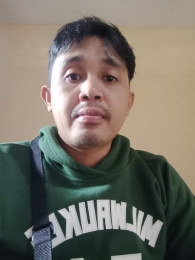 A man with dark hair and a mustache is pictured in a close-up shot. He wears a green hoodie with the text "MILWAUKEE" printed in white. A dark-colored strap crosses his upper body. The background is a plain, neutral color, drawing focus to the man's face and expression.