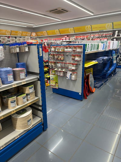 This image shows a retail store, likely a hardware or home improvement store, located in Polígon Industrial De Calvià, Spain. The scene is an aisle with shelves displaying various products. On the left side, yellow and white signage indicates product categories such as "ROPA LABORAL" (workwear), "SCREWS & FIXINGS", "BEFESTIGUNGS MATERIAL" (fastening material), "PAINTS COMPLEMENTS" (paint complements), and "ACCESORIES" (accessories). Below these signs, smaller shelves are stocked with items like screws, fasteners, and paint-related supplies.
To the right, more shelves are filled with a variety of items, including rolls of rope or cord in different colors and materials, such as natural fiber and blue plastic. Some of these rolls have "ROMBULL" branding. Further back, there are blue metal shelves holding a collection of what appear to be traffic or safety signs, including one with the text "SEÑALIZACIÓN EXCEPCIONAL" (exceptional signage) and another potentially related to emergencies or meetings ("URGENCIA", "REUNION"). A large blue tarp is also visible on a shelf.
The floor is tiled with large, light blue-gray tiles, reflecting the overhead lighting. The overall impression is of a well-stocked store with a focus on DIY, construction, and safety equipment. There are no people visible in the image, and the lighting suggests it is daytime, likely during business hours. The environment appears clean and organized.