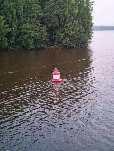A red and white buoy floats in dark, rippling water. In the background, a dense line of green trees forms a backdrop against a hazy, light-colored sky. The scene is likely on the Goritsy Reservoir, near the city of Goritsy, Russia, a body of water known for its navigation markers. The water surface shows subtle waves and reflections of the overcast sky.