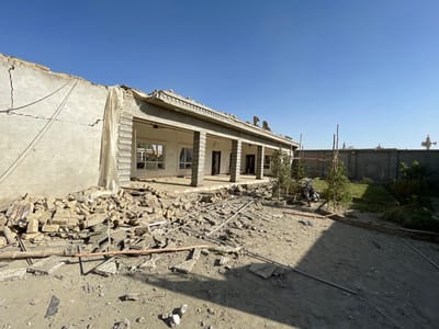 This image depicts a single-story residential or commercial structure in a state of significant disrepair, located in Spin Boldak, Afghanistan. The building, constructed from light-colored concrete or stucco, has sustained heavy damage, particularly to its roof and left-side wall, which exhibits a large crack and exposed structural elements. Portions of the roof are missing or collapsed, revealing exposed rebar and broken concrete. A fabric-like material hangs from the damaged roofline near the center.
In front of the structure, a substantial quantity of rubble, consisting of broken bricks, concrete fragments, and twisted rebar, is scattered across the ground. The building's facade features multiple pillars forming an open porch or veranda, behind which windows appear broken or entirely absent, revealing empty interior spaces.
The foreground consists of barren, dusty earth littered with additional debris, including long metal rods. Further back on the right, the ground transitions into a section of well-maintained green lawn where several small, staked trees are growing. A dark-colored object, possibly a motorcycle or utility equipment, is partially visible within this grassy area. A rudimentary scaffolding-like structure made of poles is also present near the trees.
The property is enclosed on the right by a tall, ornate concrete wall featuring decorative relief patterns. Beyond this wall, under a clear, cloudless blue sky, distant architectural elements with domed structures, possibly minarets, and a white horn-shaped loudspeaker are discernible on the horizon. No individuals are visible in the scene.
