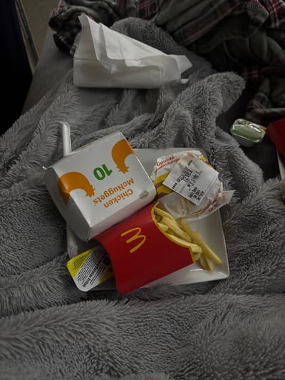 A messy scene presents McDonald's takeout on a fluffy gray blanket. A partially eaten burger, fries spilling from their red container, and a box of ten Chicken McNuggets are scattered on a white square plate. A straw and a packet of sauce sit nearby. A crumpled receipt reads "Double cheeseburger". A packet of mayonnaise, "Molho para Batatas Fritas", is also visible. A stack of napkins is on the left, near a checkered cloth.
